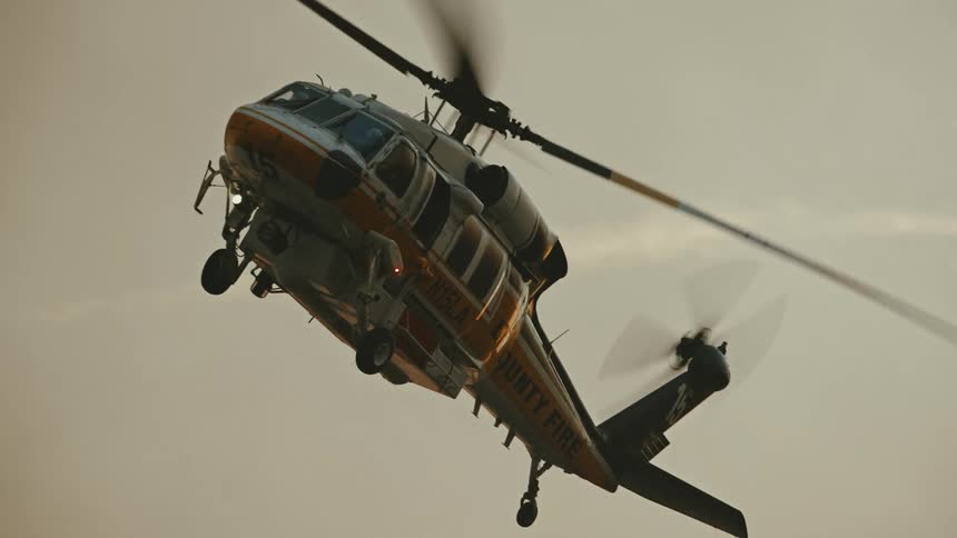 Helicopter blades turn rapidly on a yellow and white county fire helicopter ascending in front of a cloud of white smoke