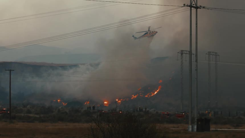 A helicopter flies above a patch of burning ground dropping water onto the flames below