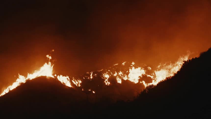 Bright orange flames on the slopes of a dark mountain ridge under a smoky sky