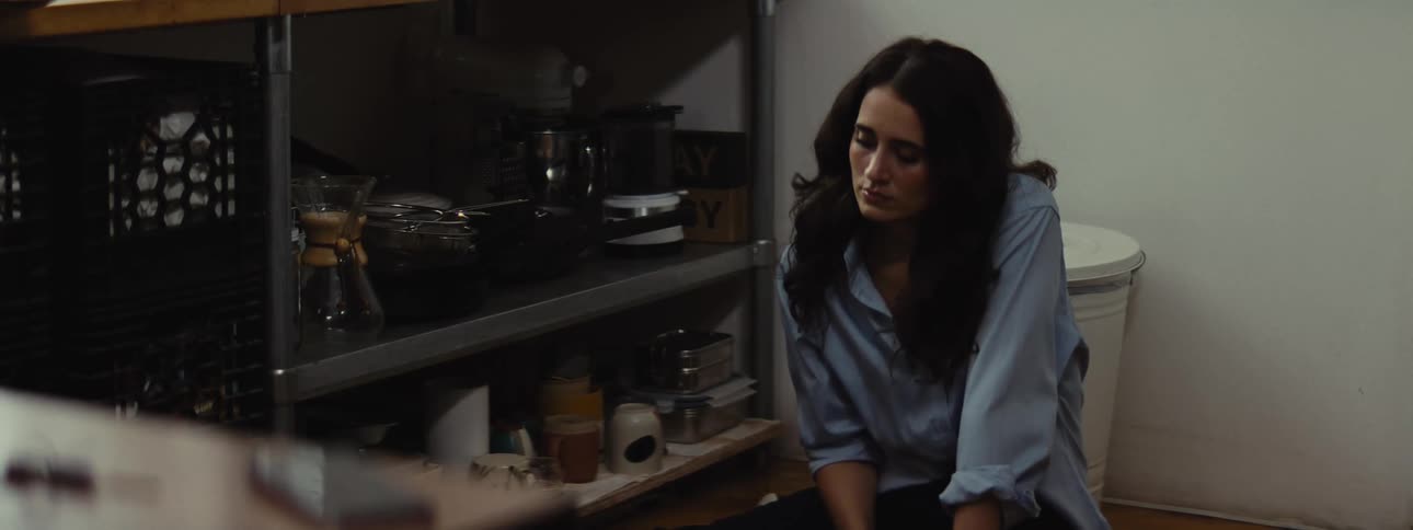 A sad woman sits on the kitchen floor looking at a phone resting on a table