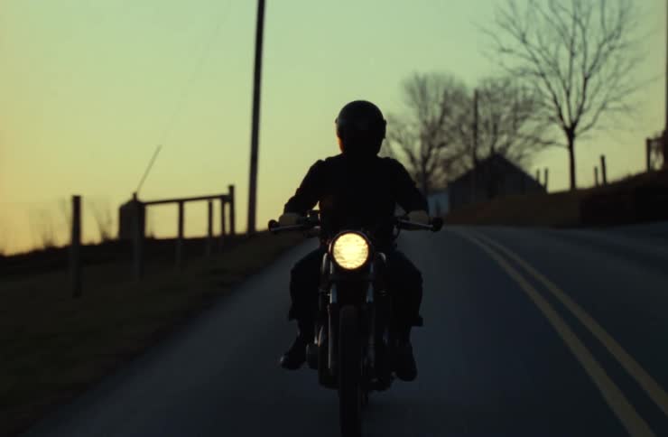 A silhouette of a motorcyclist wearing a helmet rides along a winding rural road at dusk with the motorcycles headlight glowing against the fading golden sky flanked by leafless trees wooden fences and a distant farmhouse