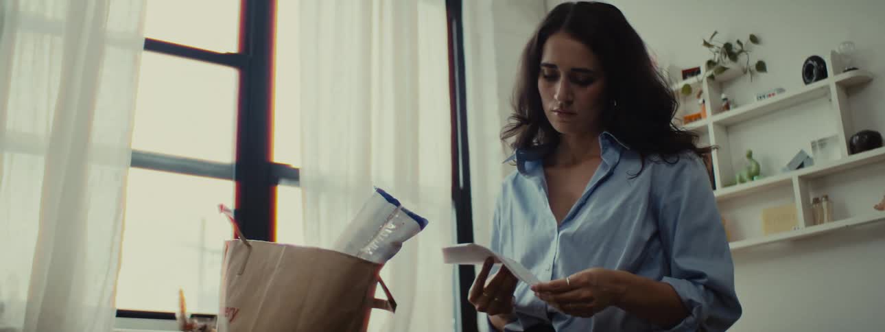 A distressed looking woman with long hair wearing a blue shirt is opening mail in the kitchen then drops the grocery bag on the floor