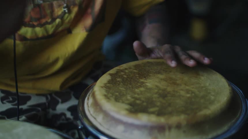 Musician in a yellow tshirt and tattooed arms plays a natural skin drum
