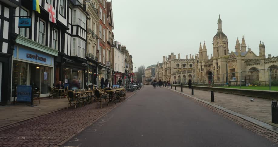An urban place with a street full of shops on one side and on the other side there is a castle with ancient architecture and several people quickly pass by