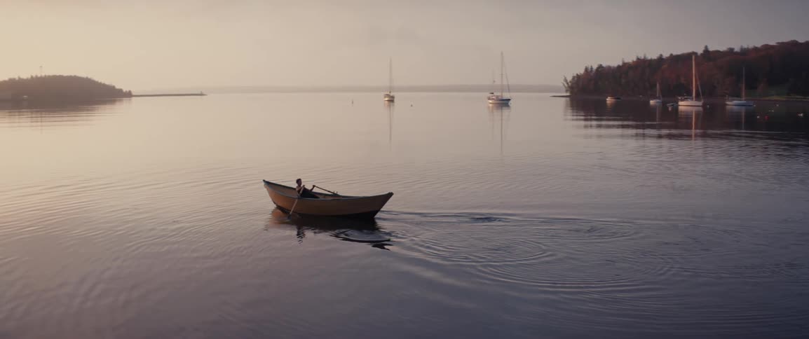 Person rowing a small boat at dawn on the calm ocean with sailboats anchored in the background