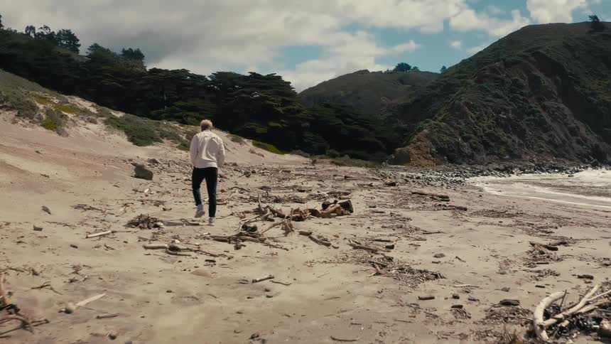 A black man is walking along a sandy beach on a partly cloudy day