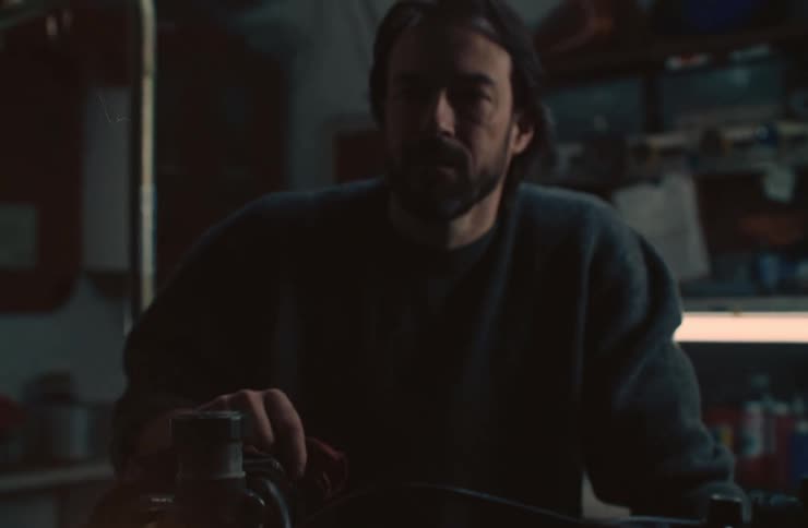The headlight of a vintage motorcycle as a bearded man wearing a sweater turns it on the warm glow of the lightbulb illuminating the inside in a dimly lit setting
