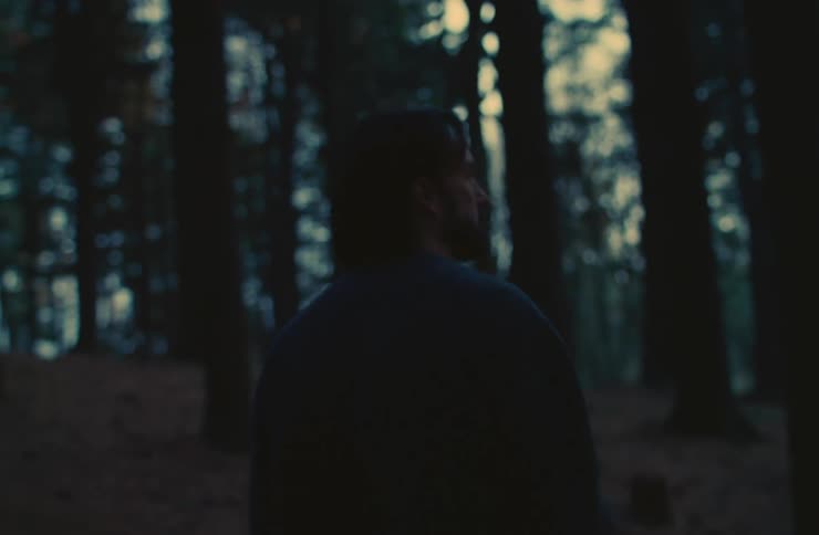 A person is walking in a dimly lit forest with tall trees in the background
