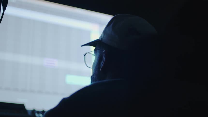 The back of the head of a man wearing a cap and glasses and looking at a bright screen in a dark room
