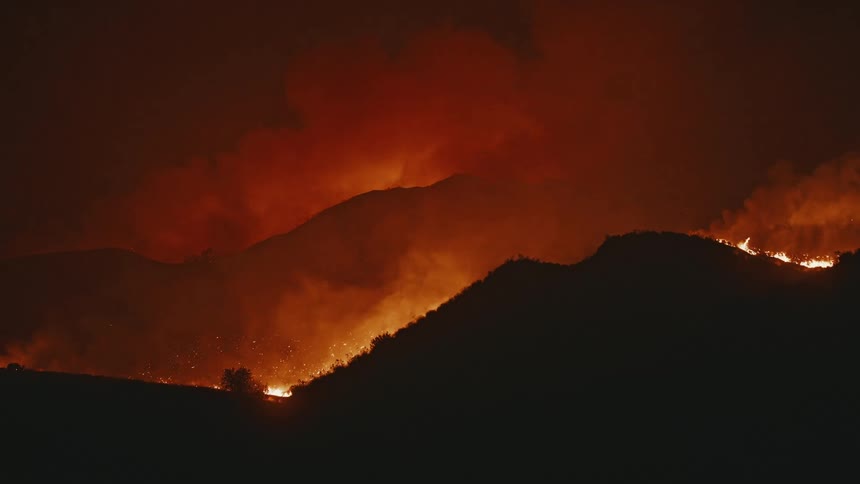 Smoke envelops the hills as a wildfires flames peek over the ridges at night