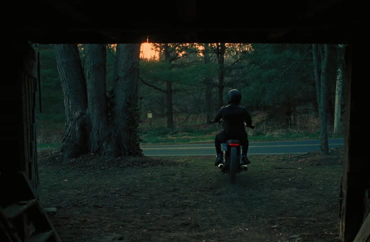 An adult rides a motorcycle out of a garage and onto a rural highway at sunset