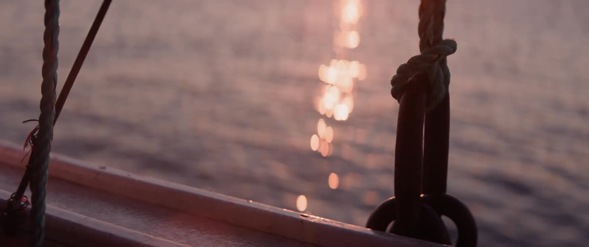 A boats rigging with ropes and metal rings softly illuminated by the warm glow of the sun reflecting off the rippling water