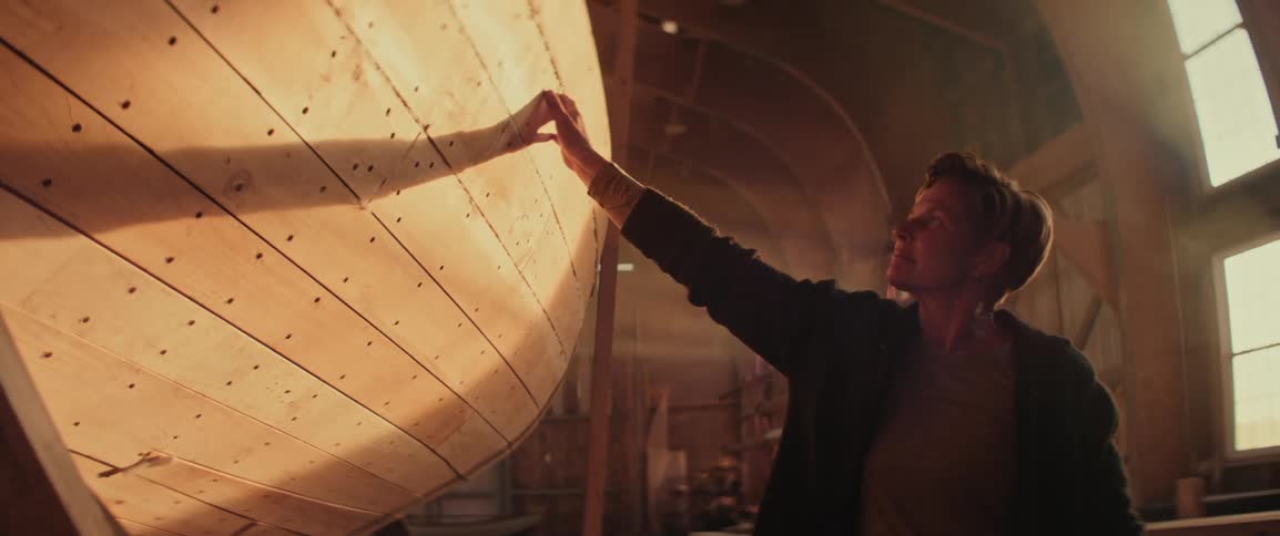 Woman with short hair and dark clothing touches the wooden structure of a boat under construction inside a workshop lit by sunlight