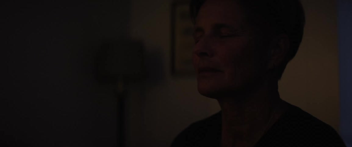 A woman sits in dim lighting with her eyes closed appearing deep in thought or meditation as light moves across the room