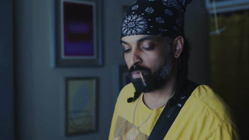A musician wearing a bandana smokes a handmade cigarette while playing music at home