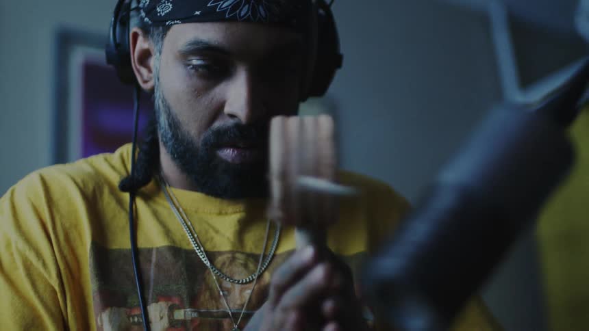 Bearded man wearing a black bandana headphones and a yellow tshirt holds a wooden percussion instrument while standing in front of a microphone