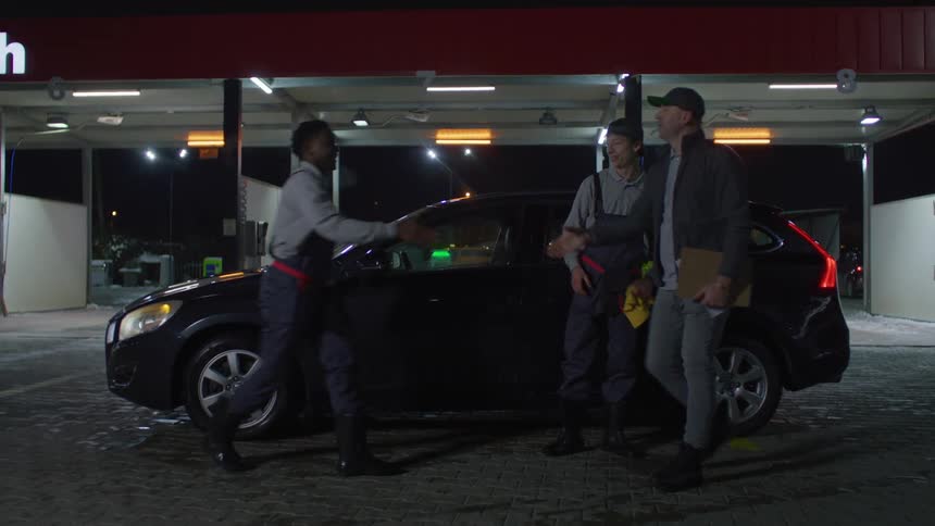 Two workers clean and detail a black car at a welllit service station at night while a third person supervises nearby holding a clipboard