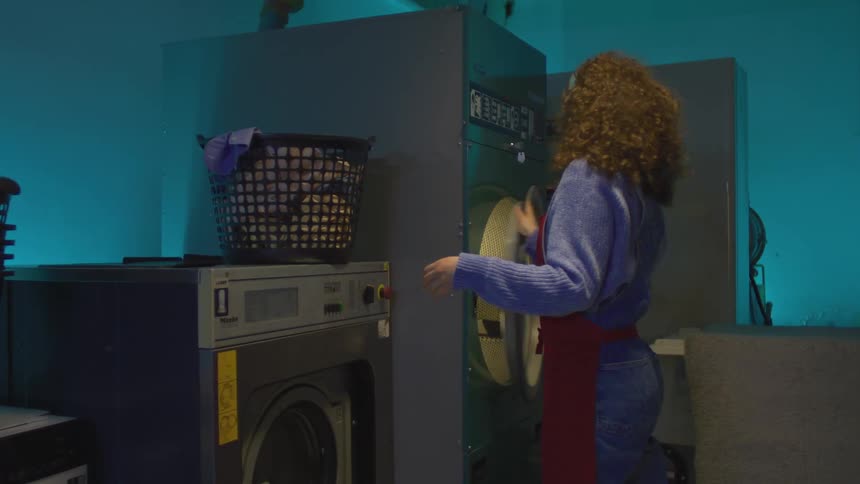 Woman with headphones takes a piece of clothing out of a washing machine and puts it in a basket