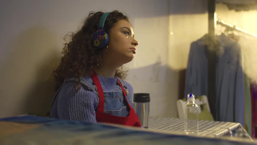 Woman with curly hair wearing colorful headphones and a blue sweater drinks water from a bottle and stands up and walks down the hallway