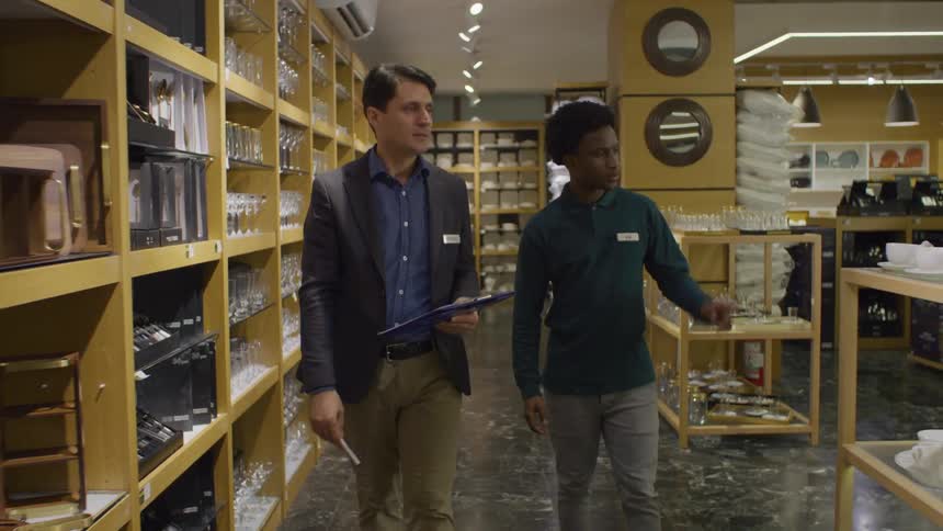 A man wearing a formal suit is walking next to a younger man and he is showing the shelf full of glass cups