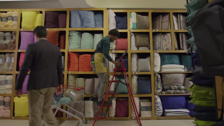 A young man placing a ladder on the floor to climb up and place a cushion on a shelf in a store