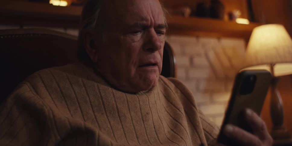 An elderly man sits in a leather armchair by a white brick fireplace holding a smartphone and laughing in astonishment