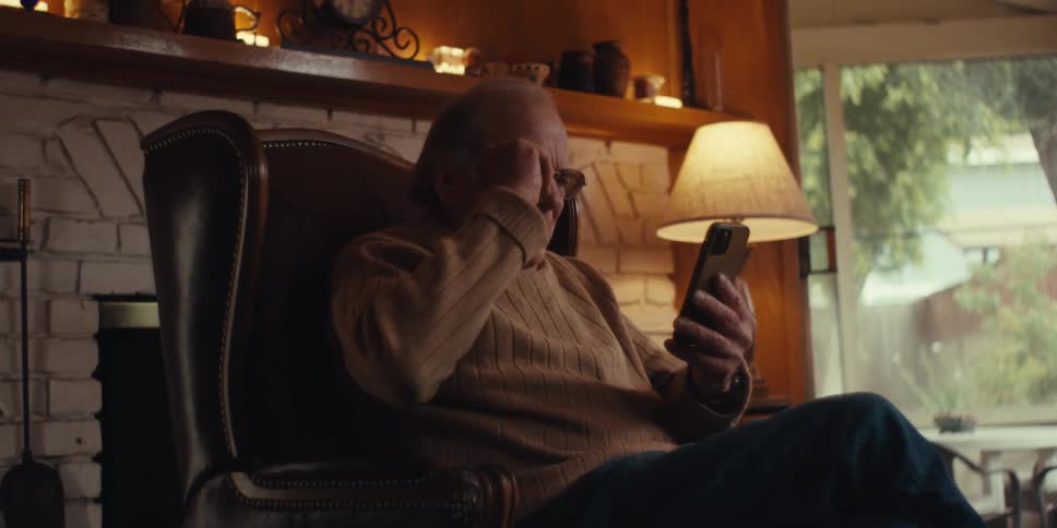 An elderly man sits in a leather armchair by a white brick fireplace holding a smartphone and laughing in astonishment