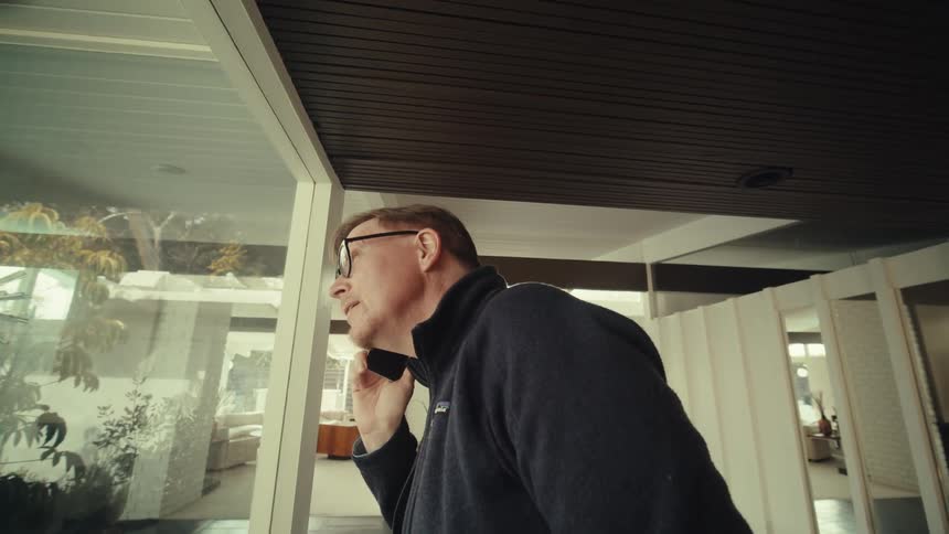 A middle aged man with glasses and a dark jacket is standing in a modern home holding a smartphone to his ear while engaged in a conversation looking out the window with a focused expression