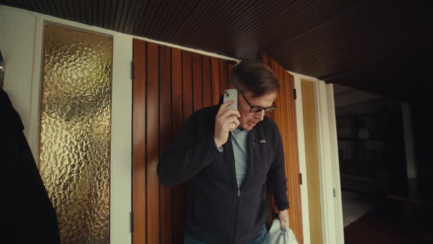 A middle aged man with glasses and a dark jacket talks on his smartphone while opening the door with a bag in his hand