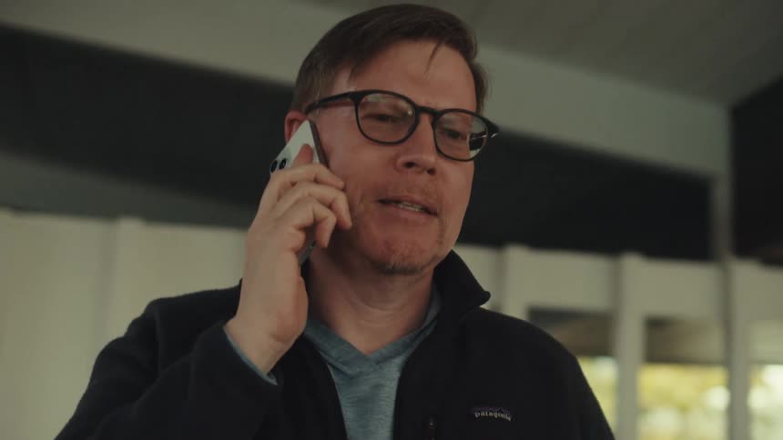 A man wearing glasses and a black jacket talks on his smartphone nodding affirmatively