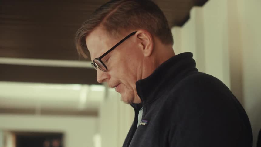 A middle aged man wearing a dark jacket and glasses uses a silver iphone by the door
