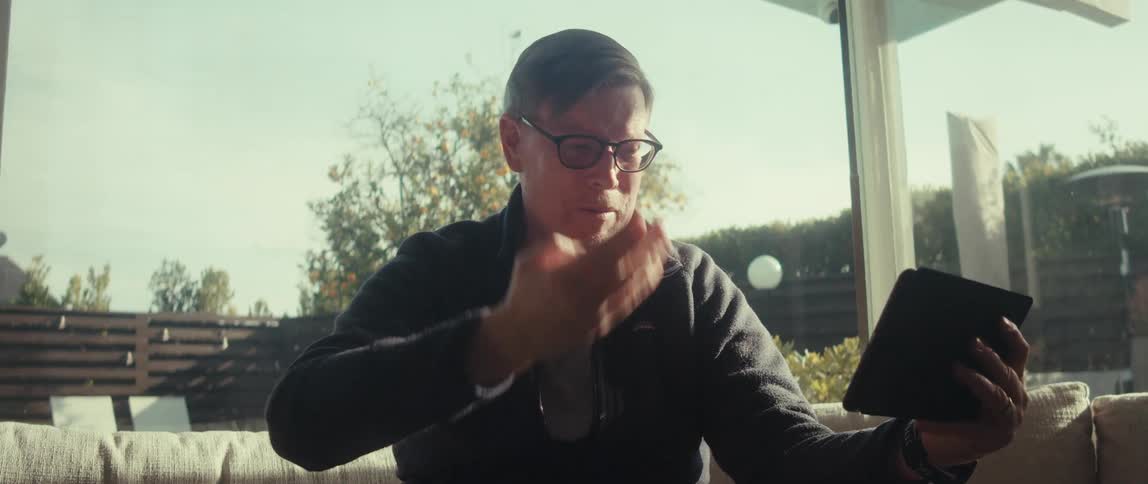 A man in glasses sits inside with a tablet looking frustrated and raising his hand
