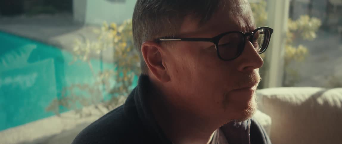 A man wearing glasses sits indoors while looking down at a tablet in his hands