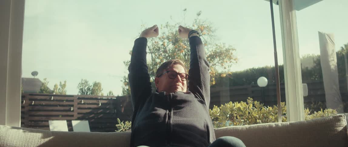Inside a sunlit room a man in glasses gestures emphatically while holding a tablet occasionally stretching out his arms