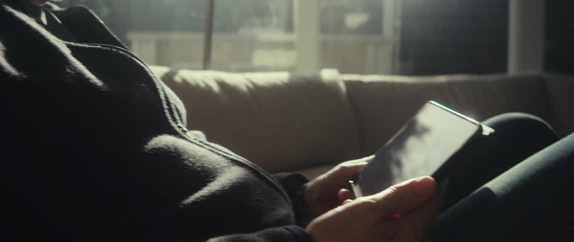 A man wearing glasses sits indoors while looking down at a tablet in his hands