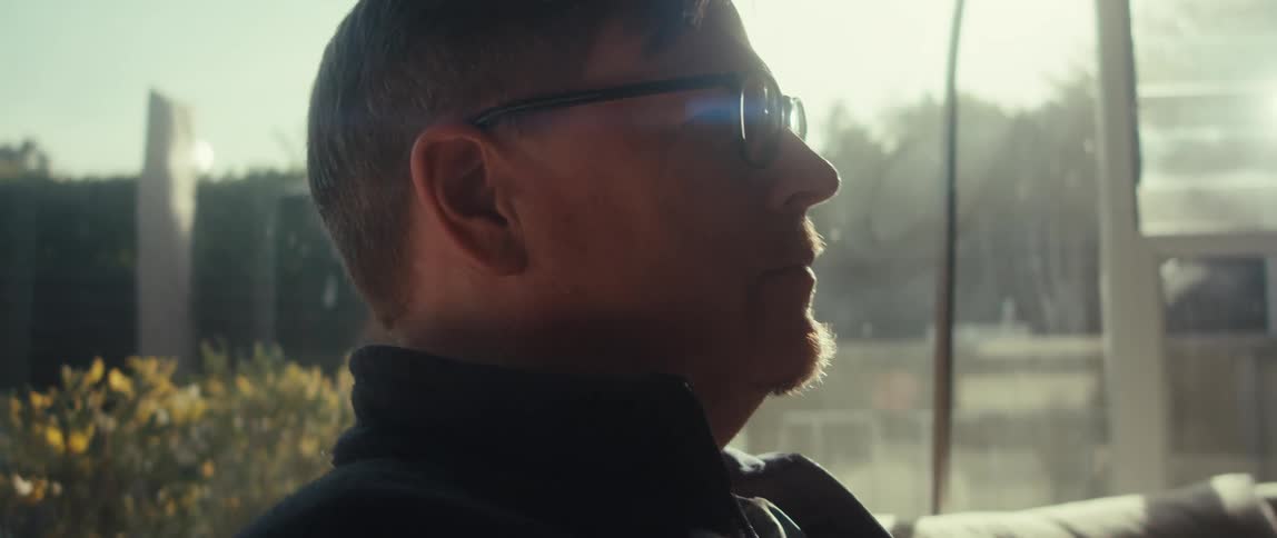 In a sun drenched room a man with glasses looks off in contemplation before focusing on his phone
