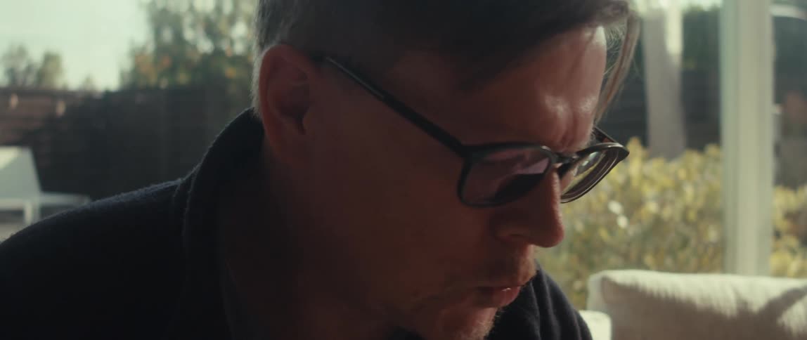 A man in glasses and a dark jacket sits on a couch and operates a tablet