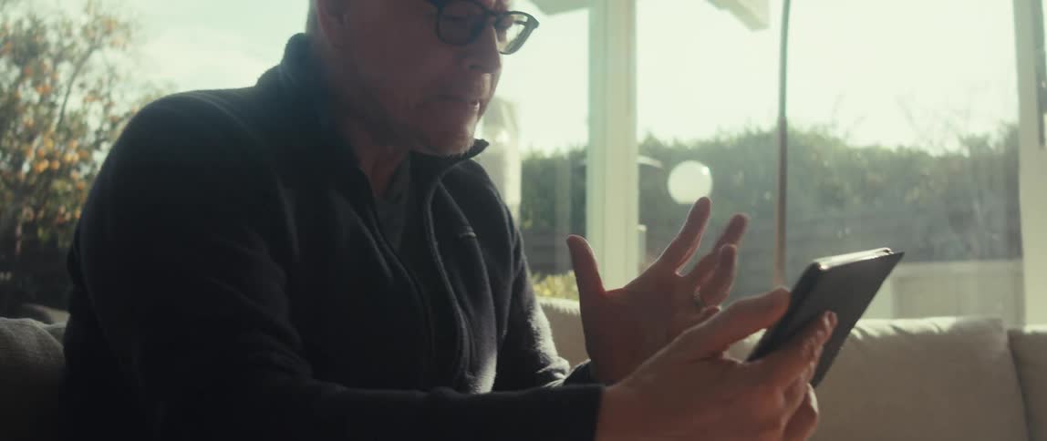 A middle aged man wearing glasses and a dark sweater sits near a large window emotionally engaged and clenching his fist while looking at a tablet