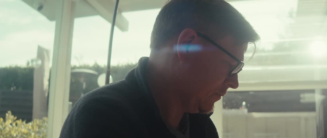 An older man with glasses sits on a couch in an enclosed patio with large windows looking down at something off screen