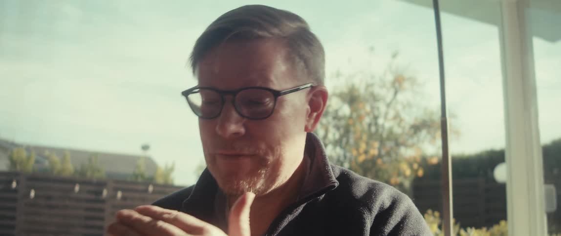 A middle aged man wearing sunglasses and a black jacket sits on the couch in the living room having a video call on a smartphone next to the swimming pool