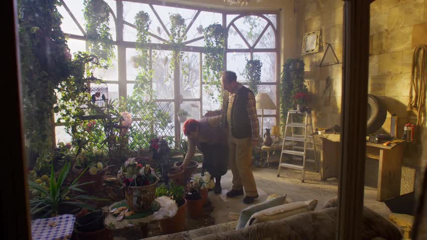 Woman watering flowers on a bench and a man comes and hugs her