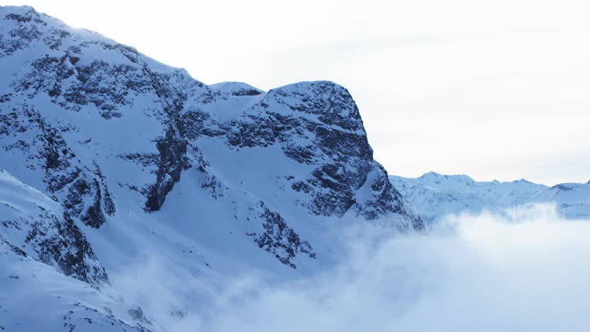 Jagged snowcovered mountain peaks rise dramatically above a swirling mist fading into a bright overcast sky