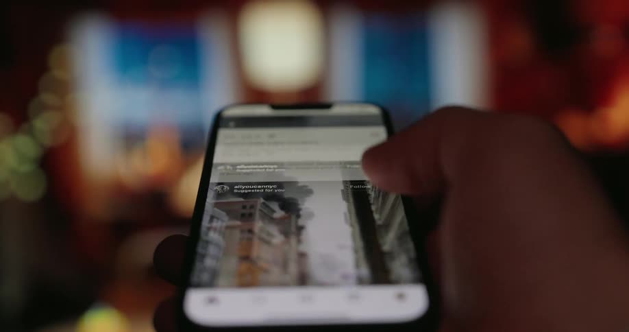 A person is shown scrolling through instagram on their iphone in a dark room with bokeh lighting in the background