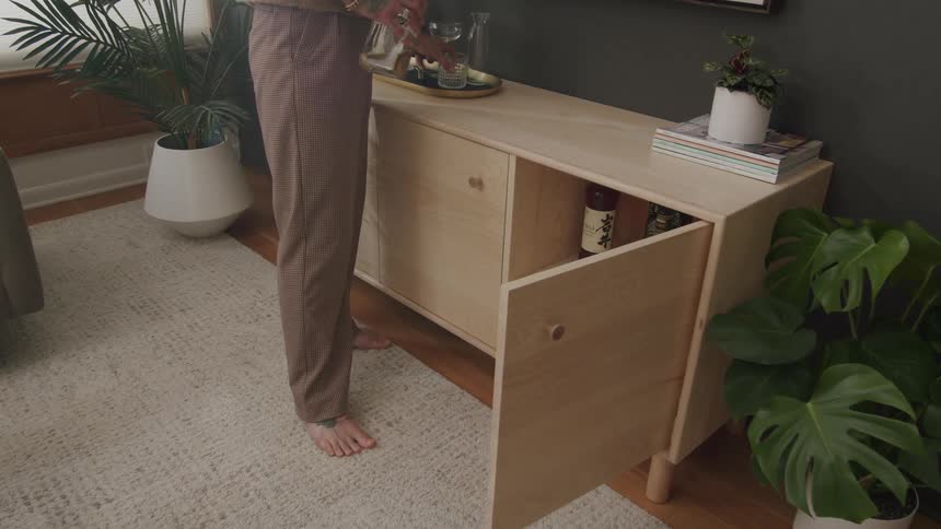 A person is serving a drink on top of a light wooden sideboard with one door open