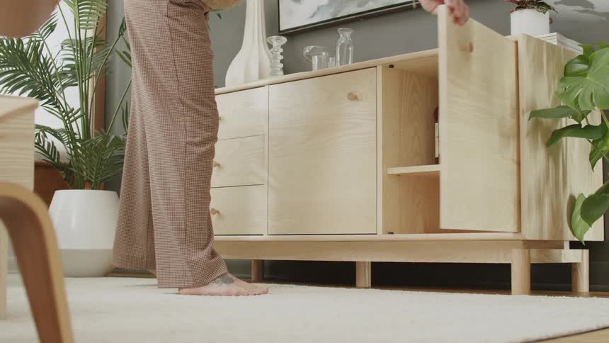 Barefoot person opens and takes a bottle of liquor from a wooden cabinet in a room decorated with plants