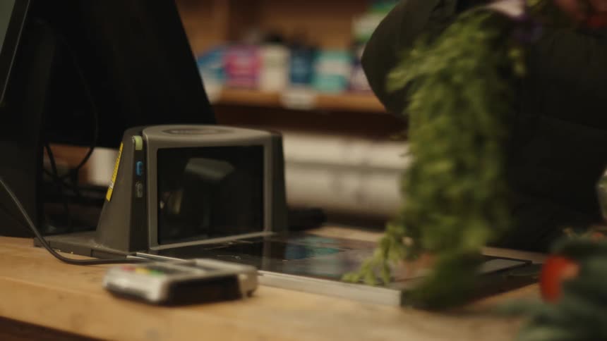 A cashier in a black jacket scans fresh produce including lettuce and carrots and leafy greens while a payment terminal sits out of focus in the foreground