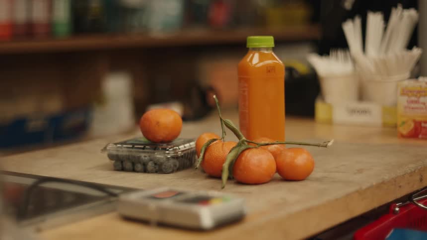 A persons hand placing some supermarket shopping on top of a wooden shelf where there are some bottles and orange fruits