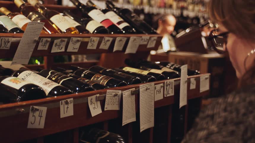 A woman is standing in front of a wine display shelf in a store examining various bottles of wine that are labeled with prices and descriptions