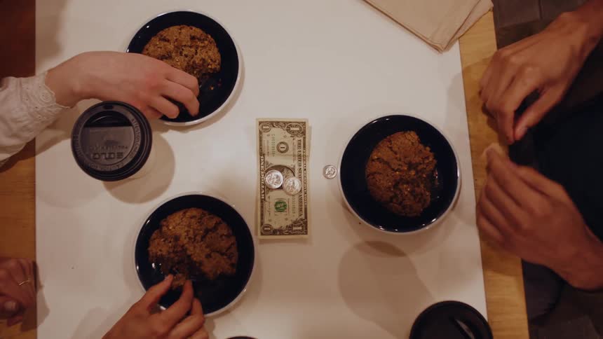 Three cookies on black plates sit on a white table accompanied by two cups of coffee and a dollar bill with some coins in the center