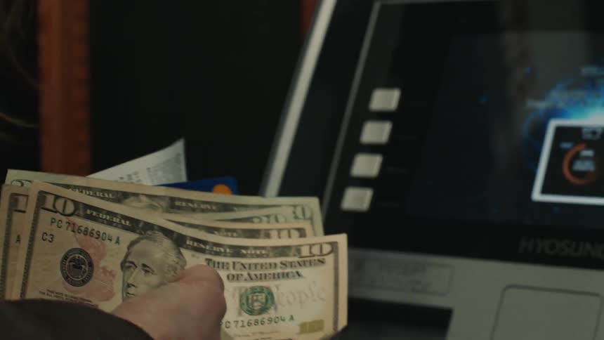 A person is holding several us dollar bills and a credit card in front of an atm machine